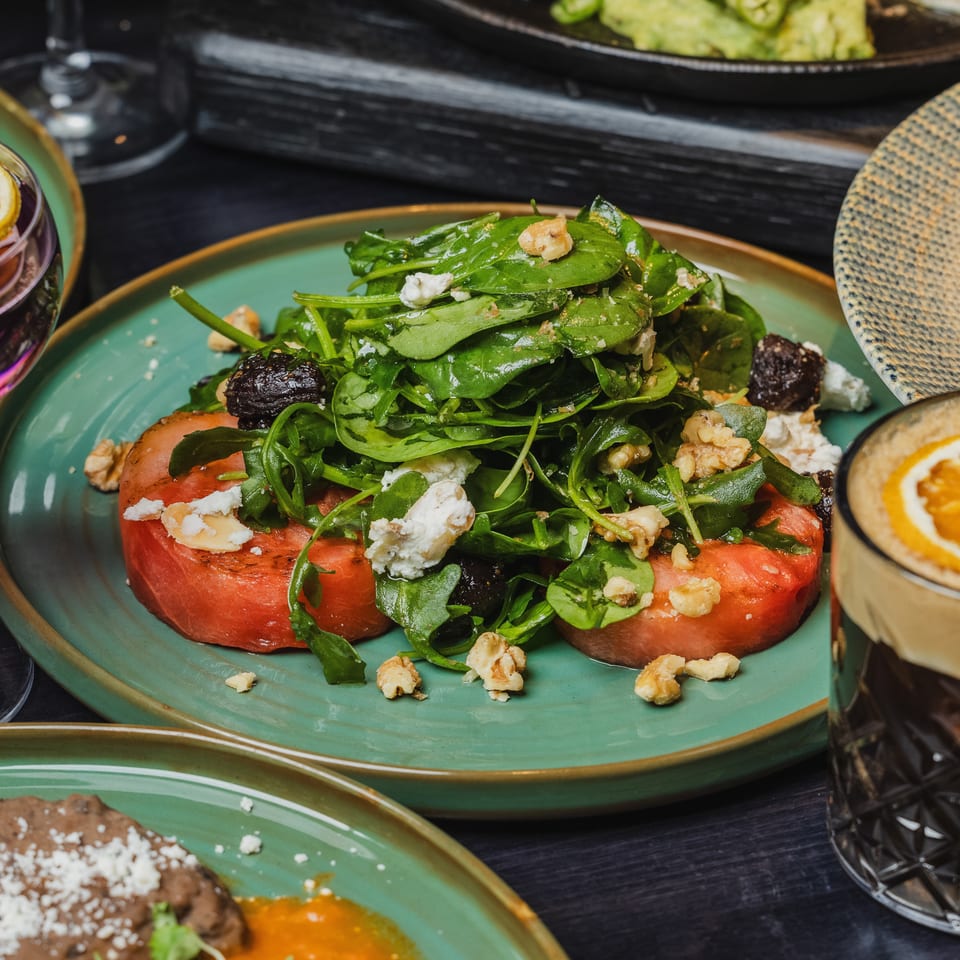 A vibrant salad with spinach, walnuts, and feta on tomato slices, alongside drinks and other dishes.