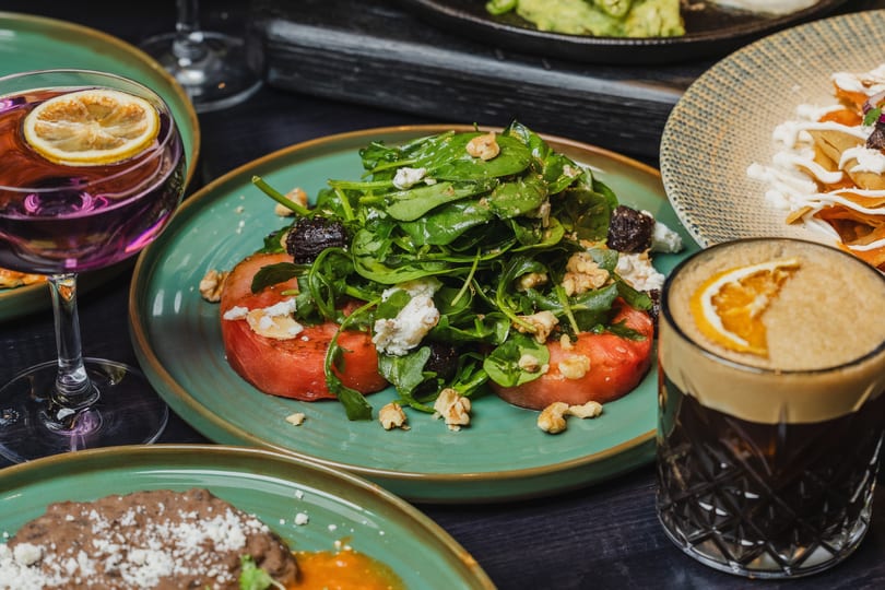 A vibrant salad with spinach, walnuts, and feta on tomato slices, alongside drinks and other dishes.
