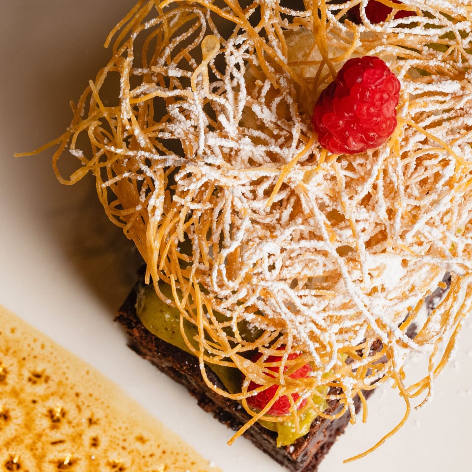 Chocolate dessert with green cream, raspberries, and a crispy sugar nest, dusted with powdered sugar.