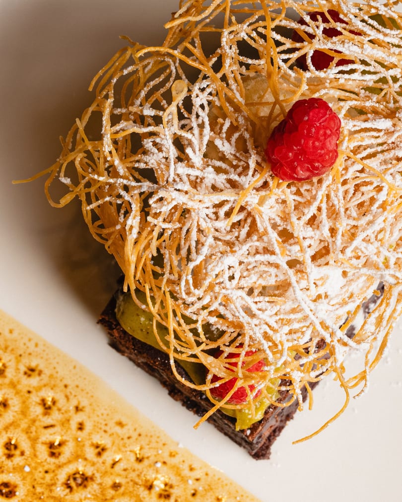 Chocolate dessert with green cream, raspberries, and a crispy sugar nest, dusted with powdered sugar.