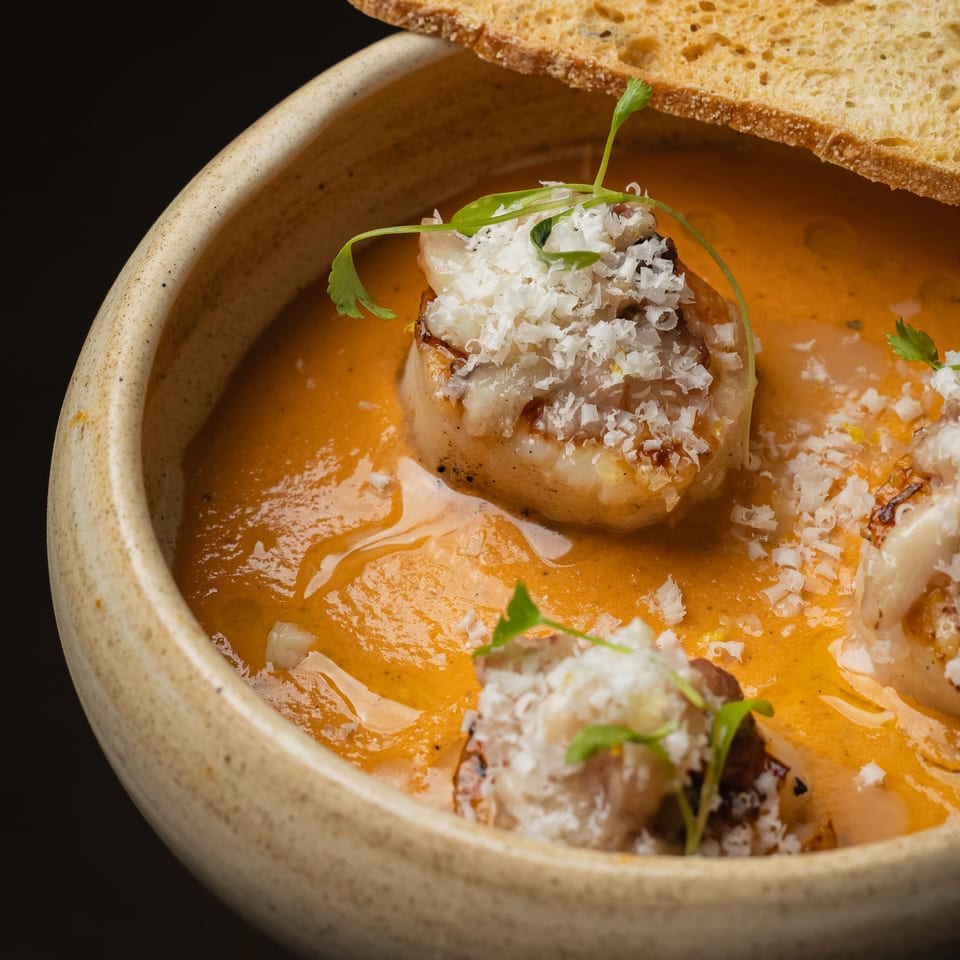 Close-up of seared scallops in orange sauce, garnished with grated cheese and microgreens, with toasted bread.