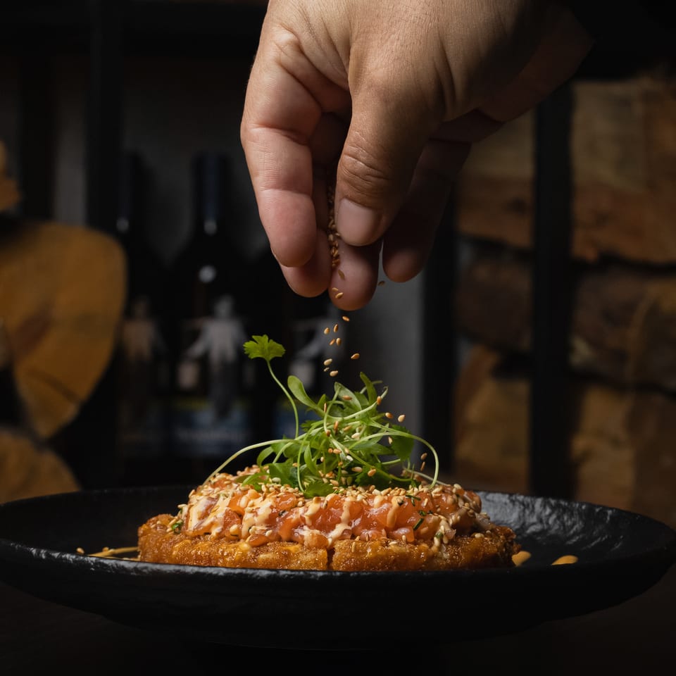 A hand sprinkles sesame seeds over a salmon dish with greens on a black plate.