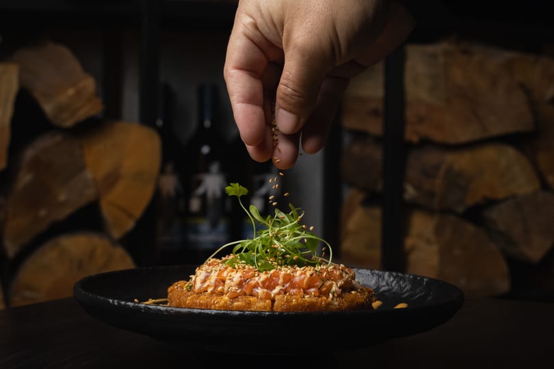 A hand sprinkles sesame seeds over a salmon dish with greens on a black plate.