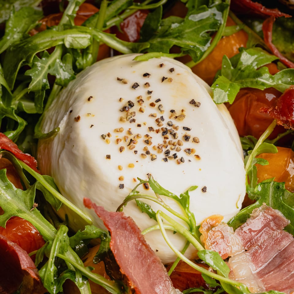 Close-up of a burrata cheese ball topped with pepper, surrounded by arugula, prosciutto, and tomatoes.