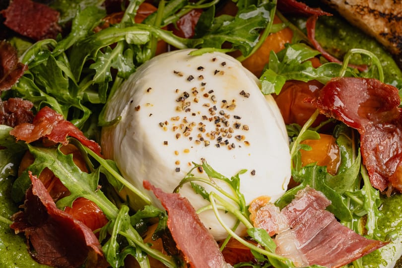 Close-up of a burrata cheese ball topped with pepper, surrounded by arugula, prosciutto, and tomatoes.