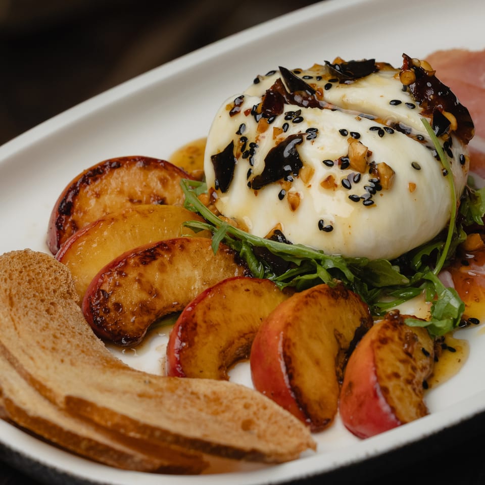 Burrata cheese with grilled peaches, arugula, sesame seeds, nuts, and toasted bread on a white plate.