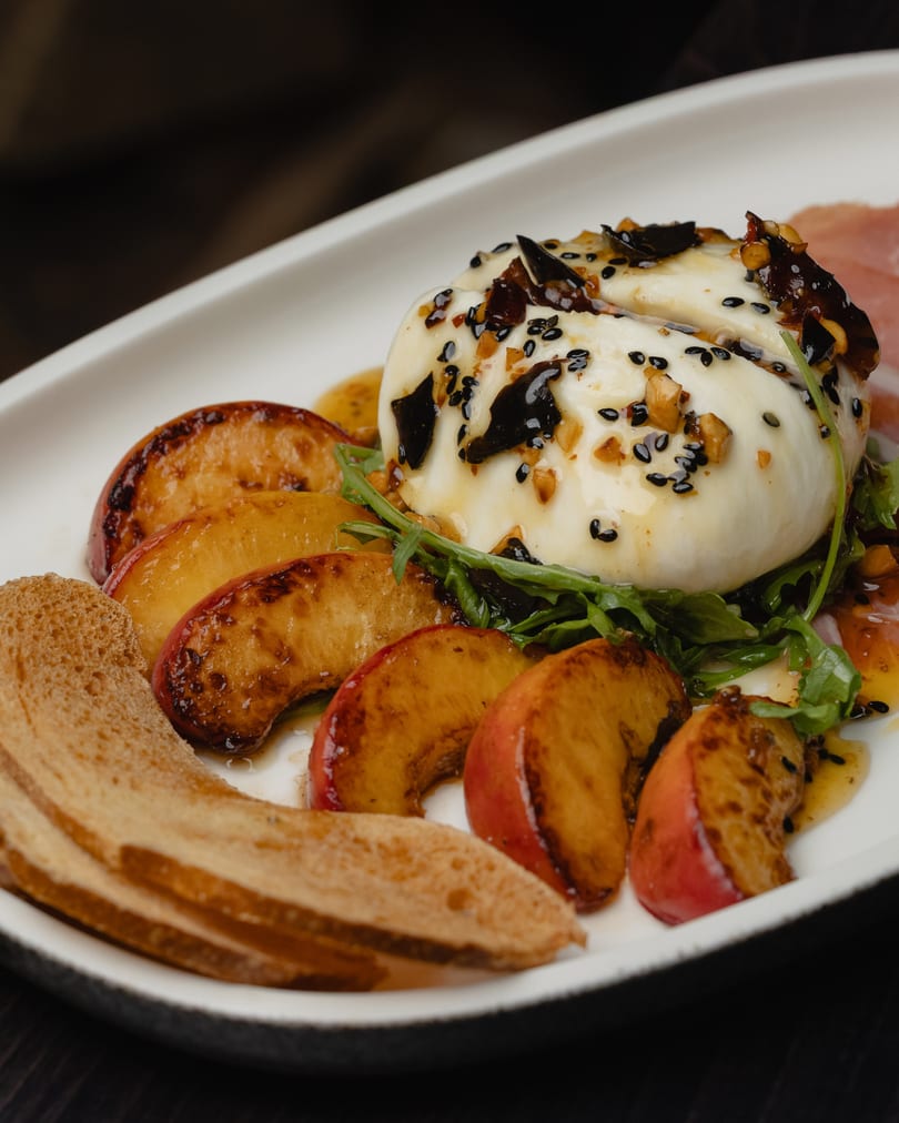 Burrata cheese with grilled peaches, arugula, sesame seeds, nuts, and toasted bread on a white plate.