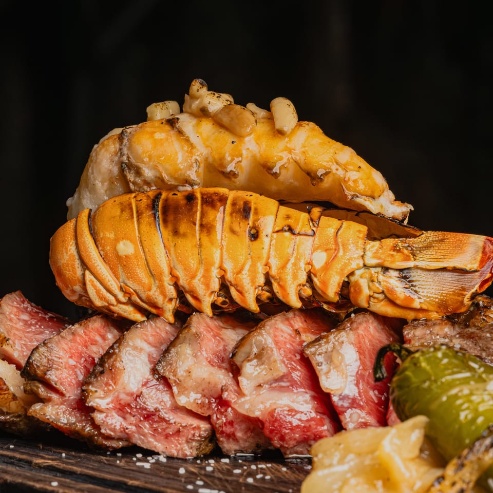 Grilled lobster tails and sliced steak with green peppers and onions on a dark wooden board.