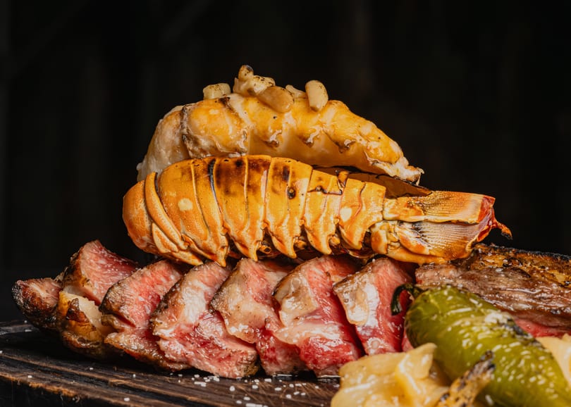 Grilled lobster tails and sliced steak with green peppers and onions on a dark wooden board.