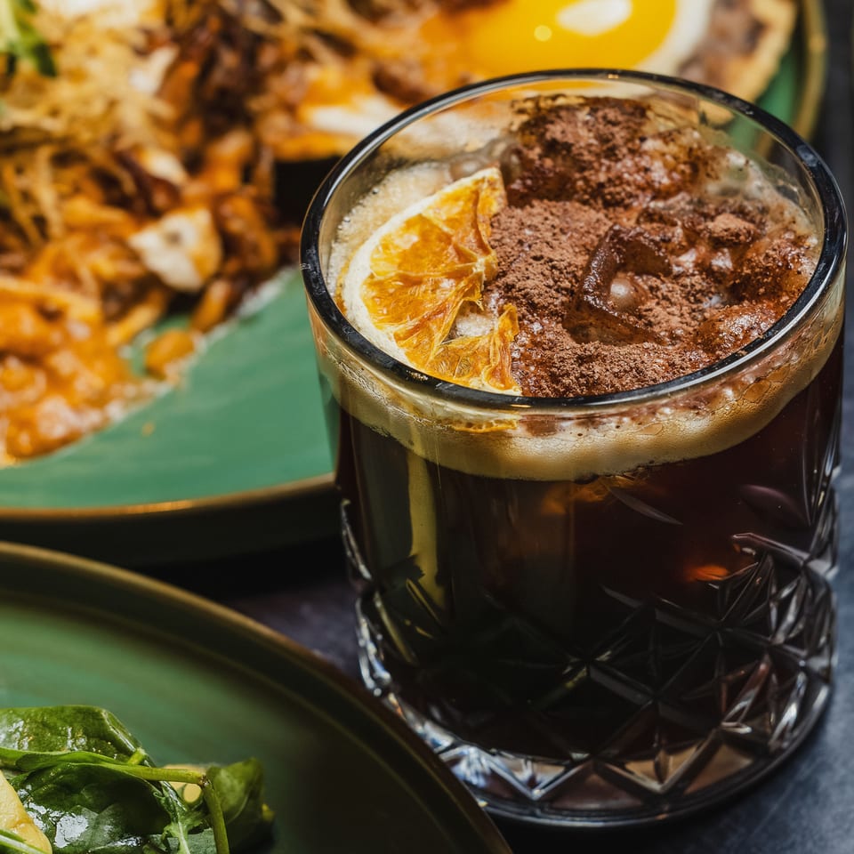 Close-up of a dark cocktail with an orange slice and cocoa powder, with food in the background.