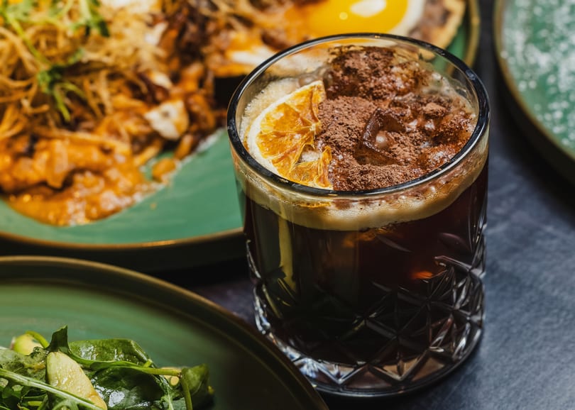 Close-up of a dark cocktail with an orange slice and cocoa powder, with food in the background.