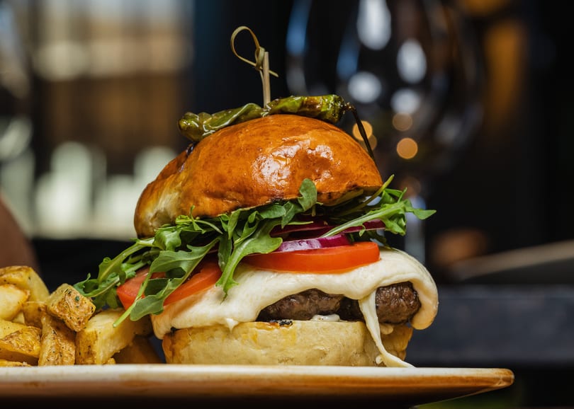 Gourmet burger with melted cheese, tomato, red onion, arugula, and a grilled pepper on top.