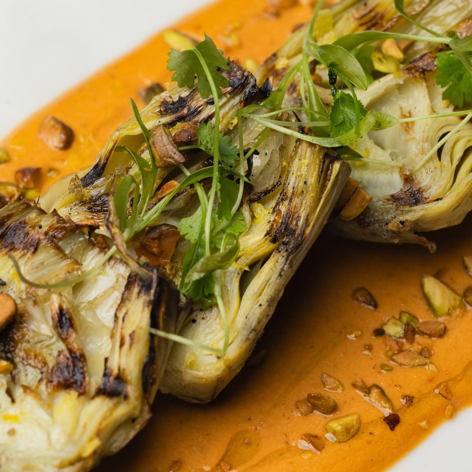 Grilled artichoke halves with fresh herbs and chopped nuts, served on a vibrant orange sauce.