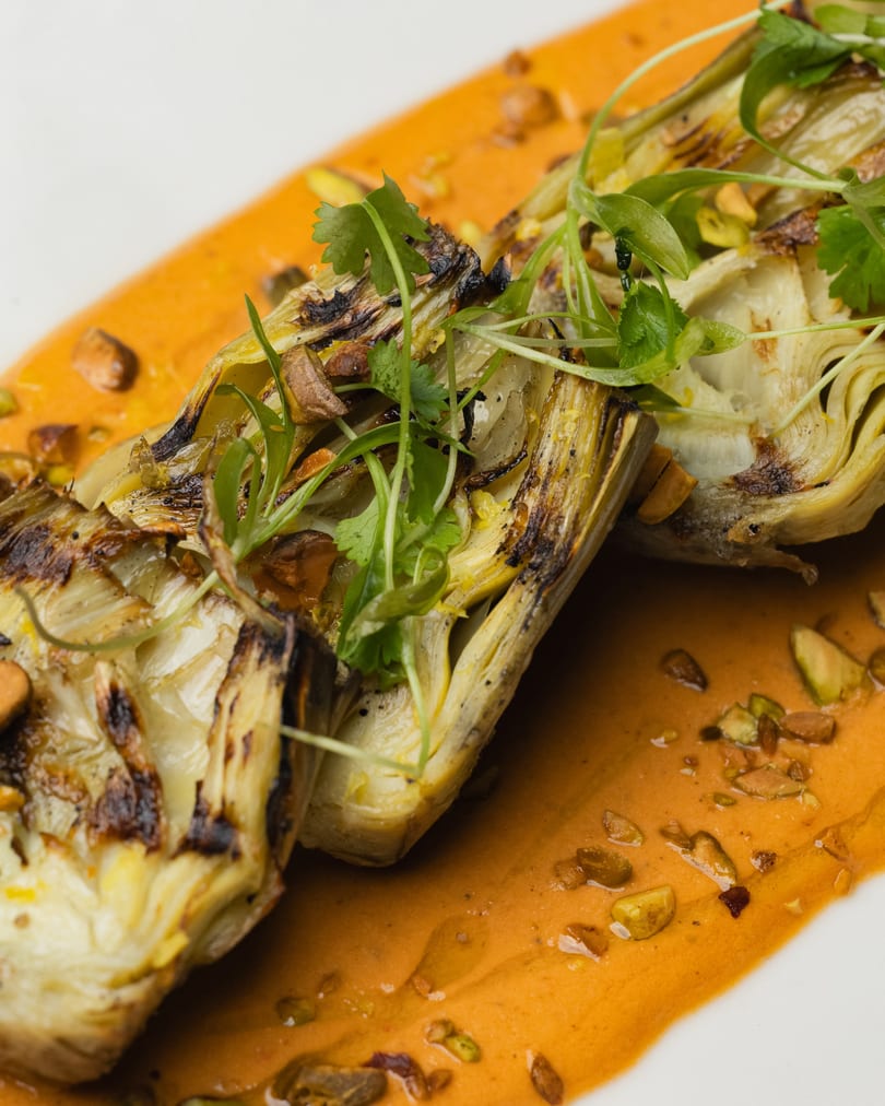 Grilled artichoke halves with fresh herbs and chopped nuts, served on a vibrant orange sauce.