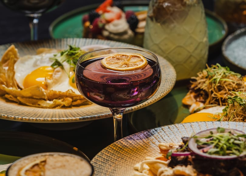 A vibrant table spread with a purple cocktail, fried eggs, and other dishes.