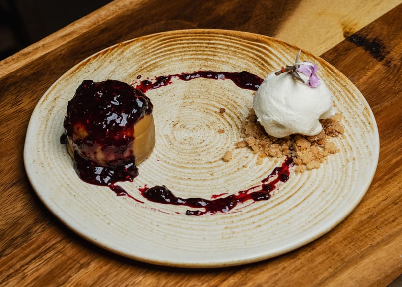 A dessert with berry compote, a scoop of ice cream, and crumble on a textured plate.