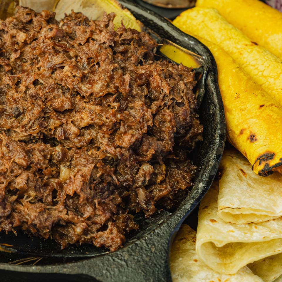 Shredded meat in a cast iron skillet, with yellow and white tortillas and red onions.