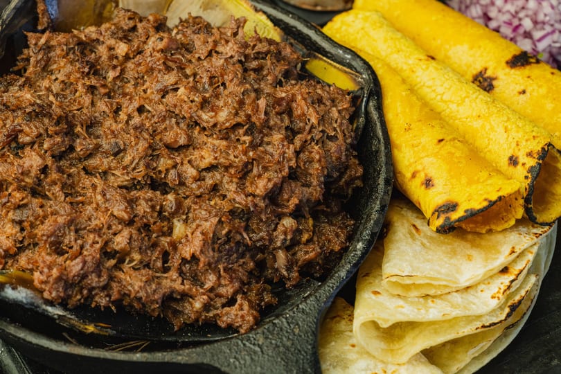 Shredded meat in a cast iron skillet, with yellow and white tortillas and red onions.