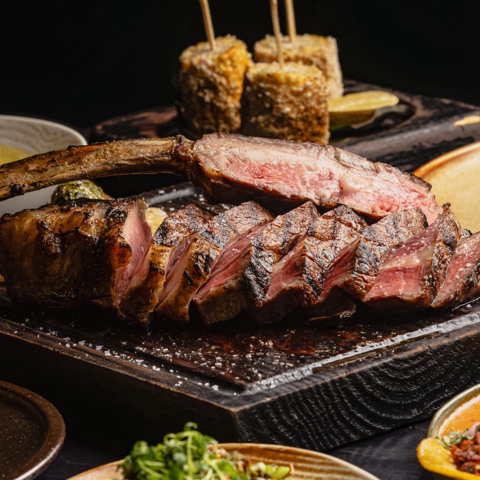 Sliced tomahawk steak on a wooden board, surrounded by various side dishes like corn and tacos.