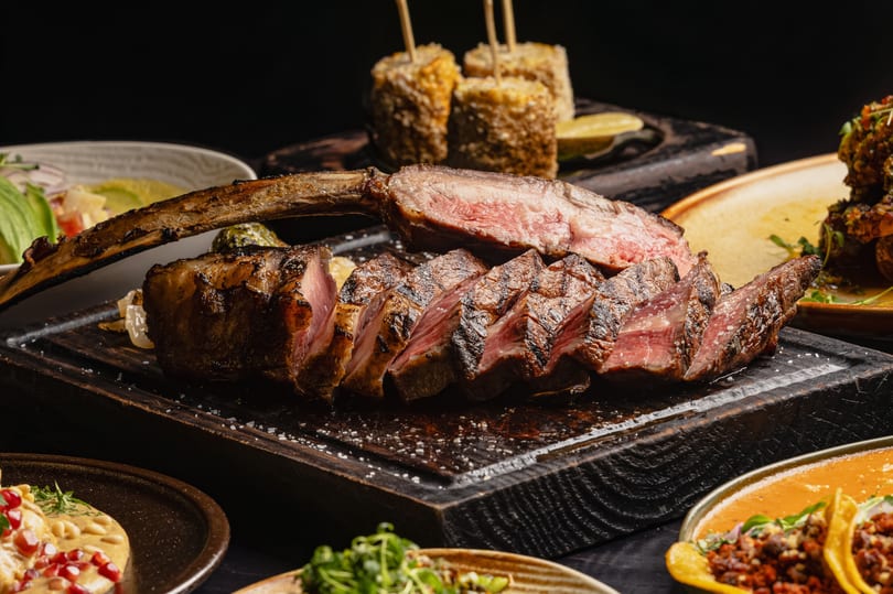 Sliced tomahawk steak on a wooden board, surrounded by various side dishes like corn and tacos.