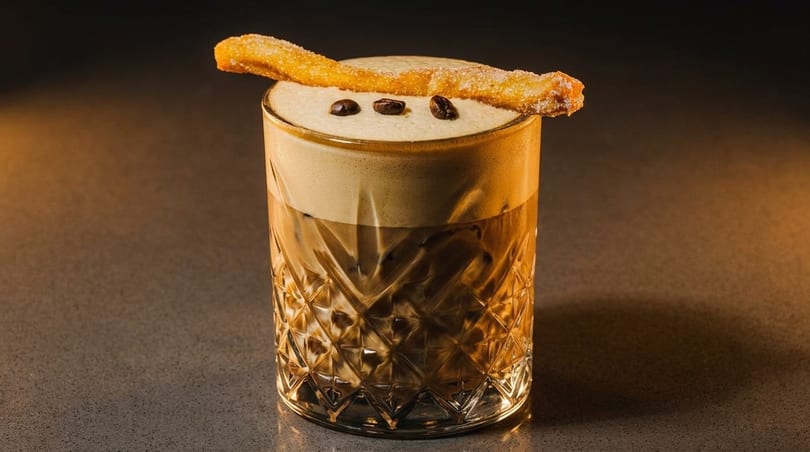 Iced coffee with foam, three coffee beans, and a churro garnish in a textured glass.