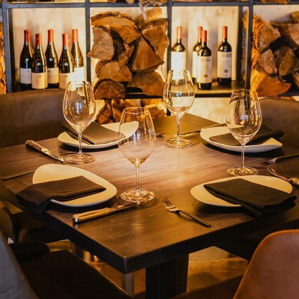 Set dining table with wine glasses and plates, against a backdrop of stacked firewood and wine bottles.