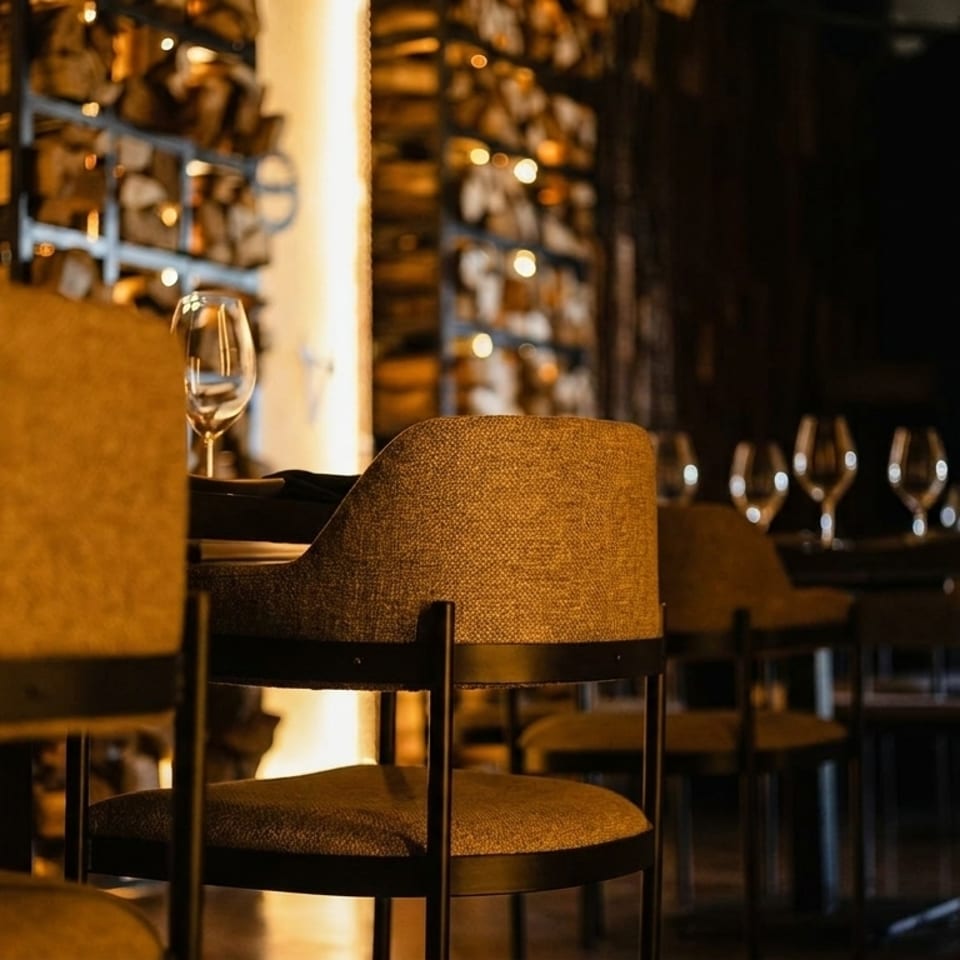 Warmly lit restaurant interior with upholstered chairs, wine glasses, and stacked firewood in the background.
